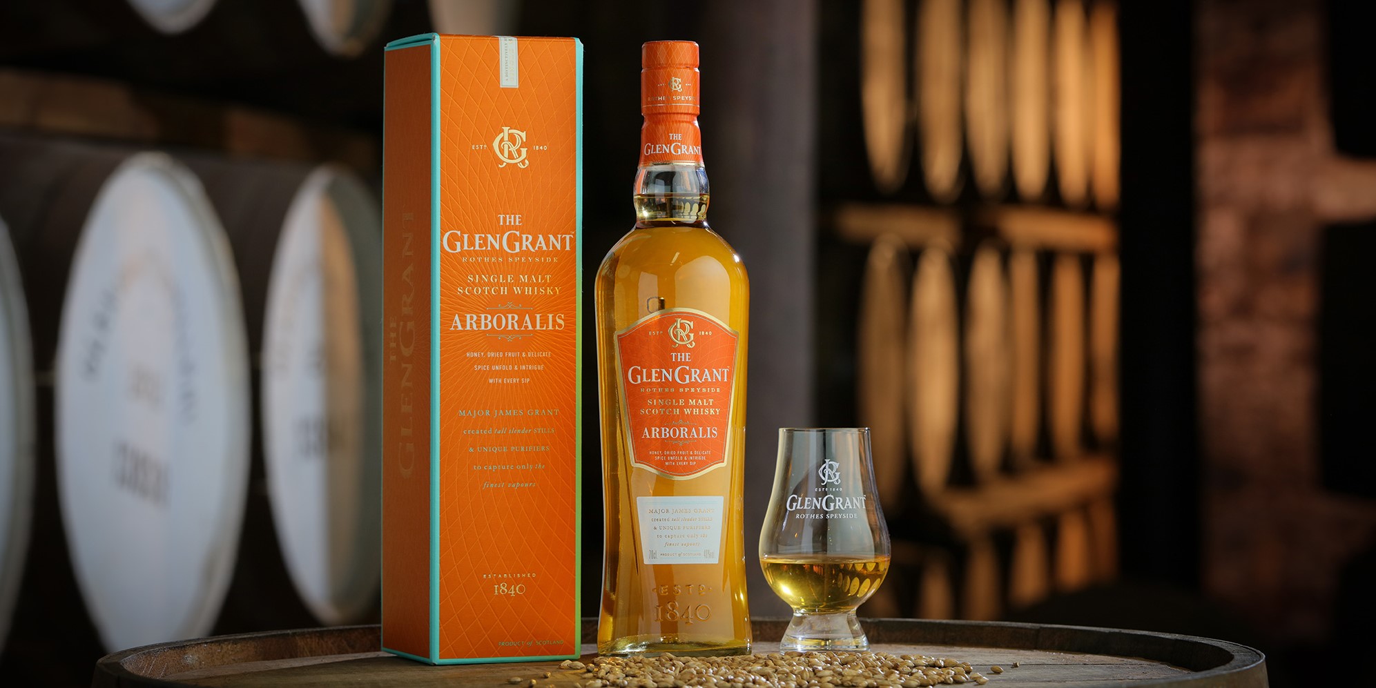 A bottle of The Glen Grant Arboralis single malt Scotch whisky stands next to its matching orange box and a filled whisky glass, with barrels and a warm, rustic background. Grains are scattered on the wooden surface.