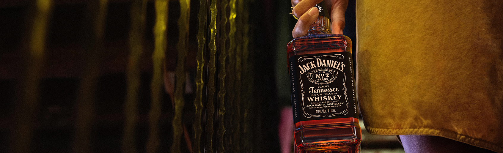A person wearing a yellow outfit holds a bottle of Jack Daniel’s Old No. 7 Tennessee Whiskey by their side, with a blurred background featuring vertical lines and warm lighting.