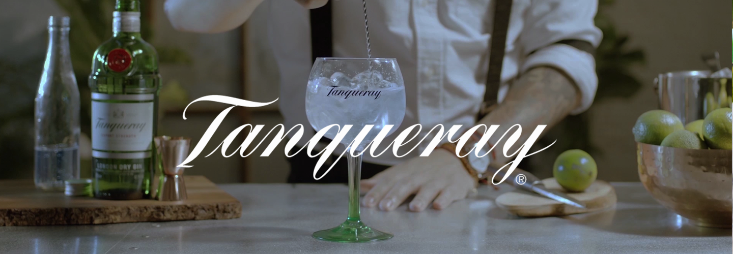 A bartender prepares a cocktail with ice in a large glass labeled Tanqueray. A Tanqueray gin bottle, limes, and bar tools are on the counter. The Tanqueray logo is prominently displayed across the image.