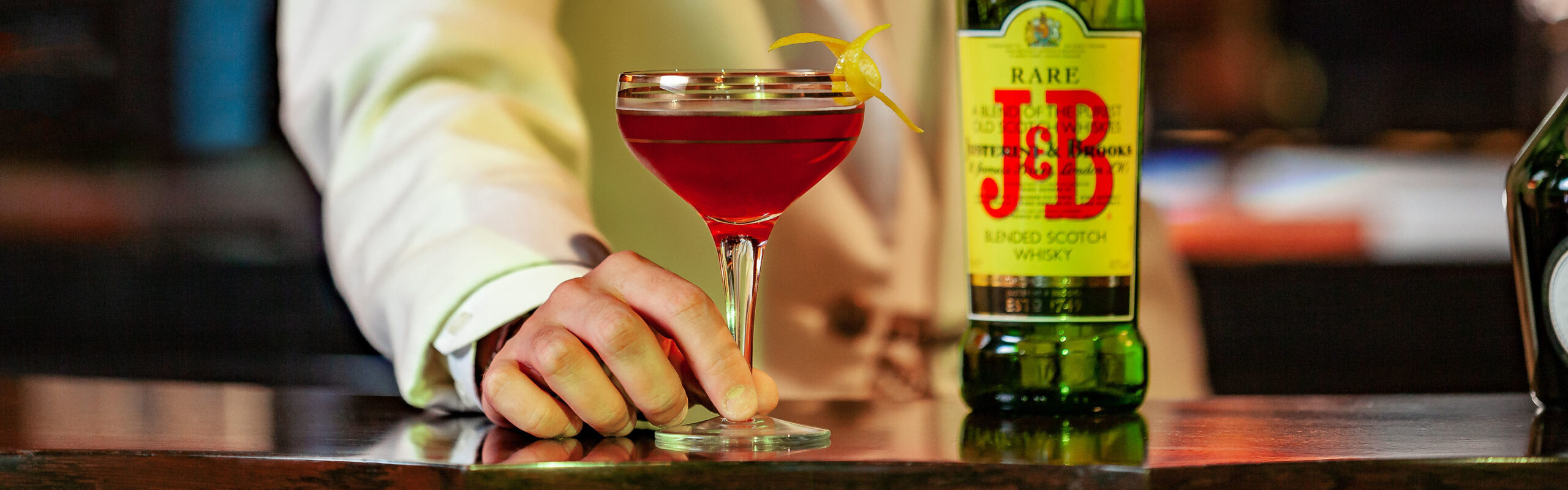 A person in a white jacket holds a cocktail in a coupe glass garnished with a lemon twist, next to a green bottle of J&B Rare Scotch Whisky on a polished bar counter.