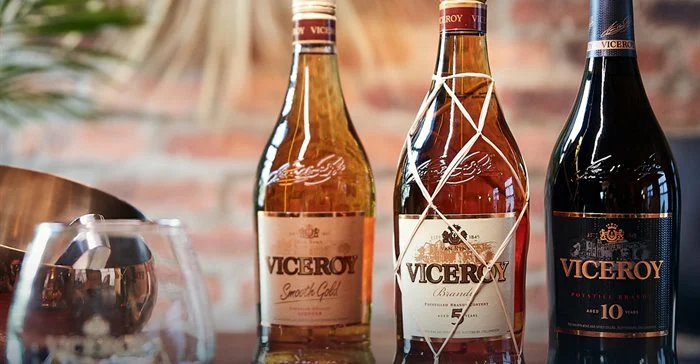 Three bottles of Viceroy brandy are displayed on a reflective surface with two glass tumblers nearby; the background is blurred with warm lighting and a hint of a plant.